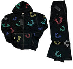 A black zip-up hoodie and matching black pants laid flat on a gray textured surface. Both pieces feature colorful embroidered horseshoe motifs in multiple neon-like colors including green, blue, red, yellow, and white.