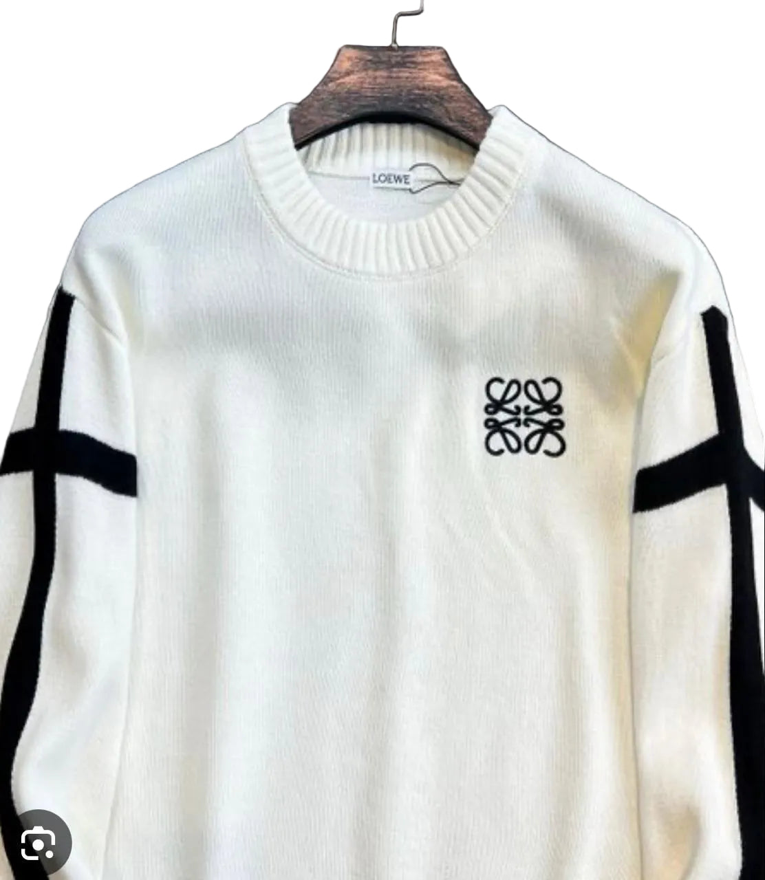 An off-white knit sweater with black stripe details on the sleeves and a black Loewe logo on the chest, hanging on a wooden hanger against a dark textured background.