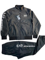 A black zip-up jacket and matching jogger pants set from Emporio Armani, laid flat on a gray textured background. The jacket features a white EA7 logo on the left chest and vertical white branding on the sleeve, while the pants display the EA7 logo and Emporio Armani text on the front.