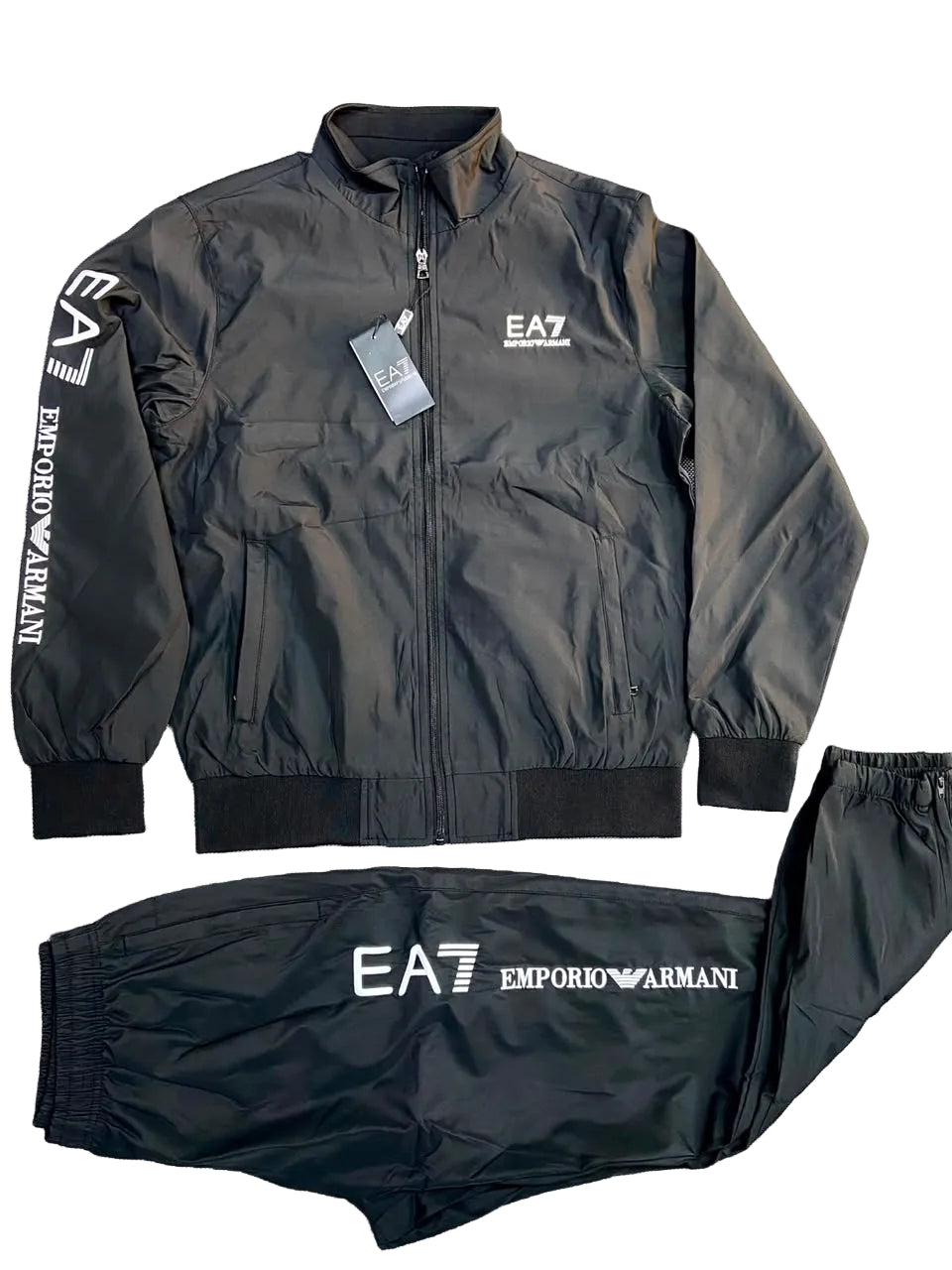 A black zip-up jacket and matching jogger pants set from Emporio Armani, laid flat on a gray textured background. The jacket features a white EA7 logo on the left chest and vertical white branding on the sleeve, while the pants display the EA7 logo and Emporio Armani text on the front.