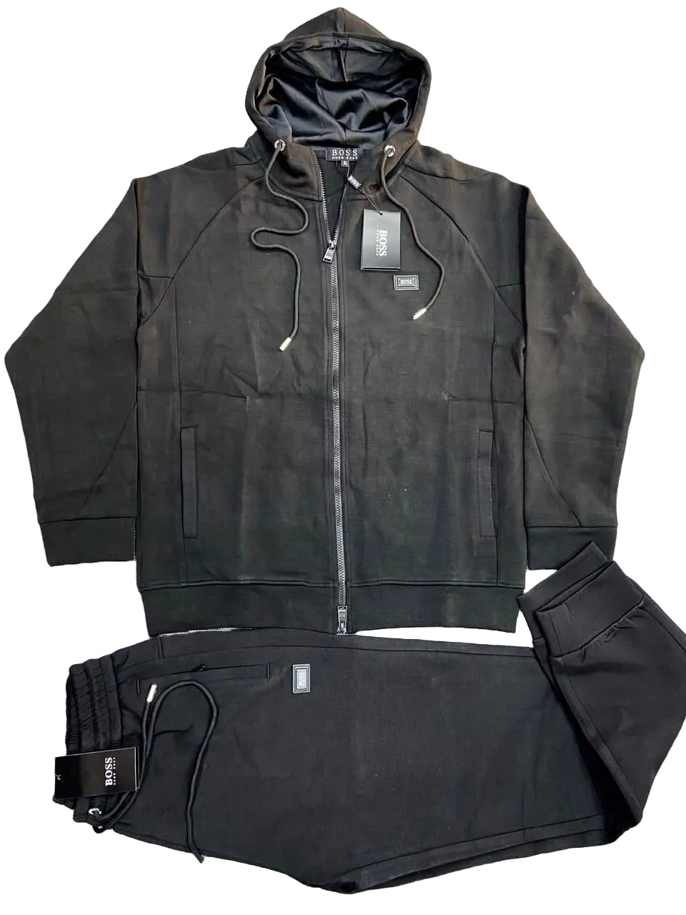 A black two-piece tracksuit laid flat on a gray textured background, featuring a zip-up hoodie with a hood and matching black jogger pants with drawstrings.
