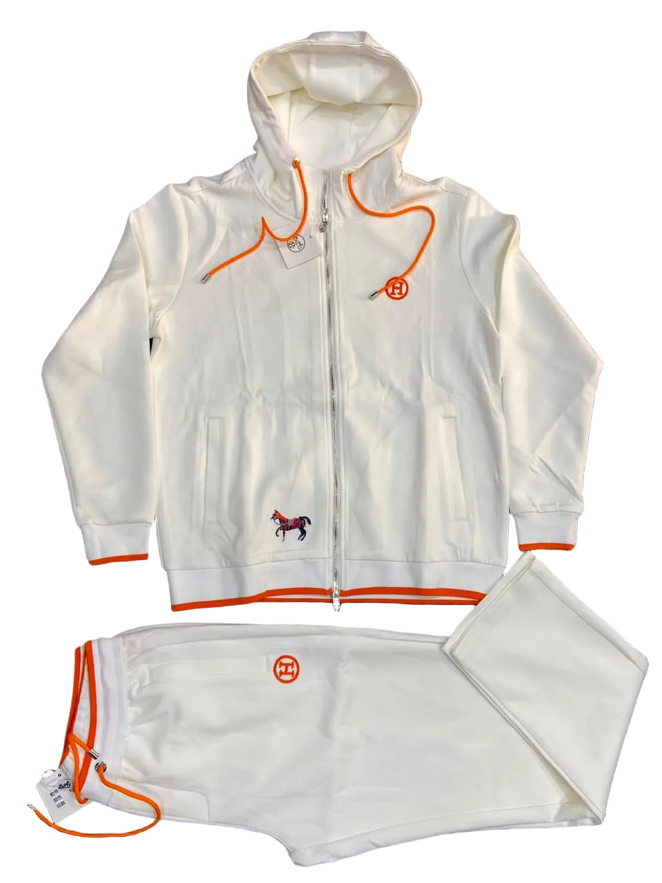 A white zip-up hoodie and matching sweatpants set laid flat on a dark gray textured background. The tracksuit features bright orange drawstrings, trim, and logo details, along with a small embroidered horse logo on the chest.