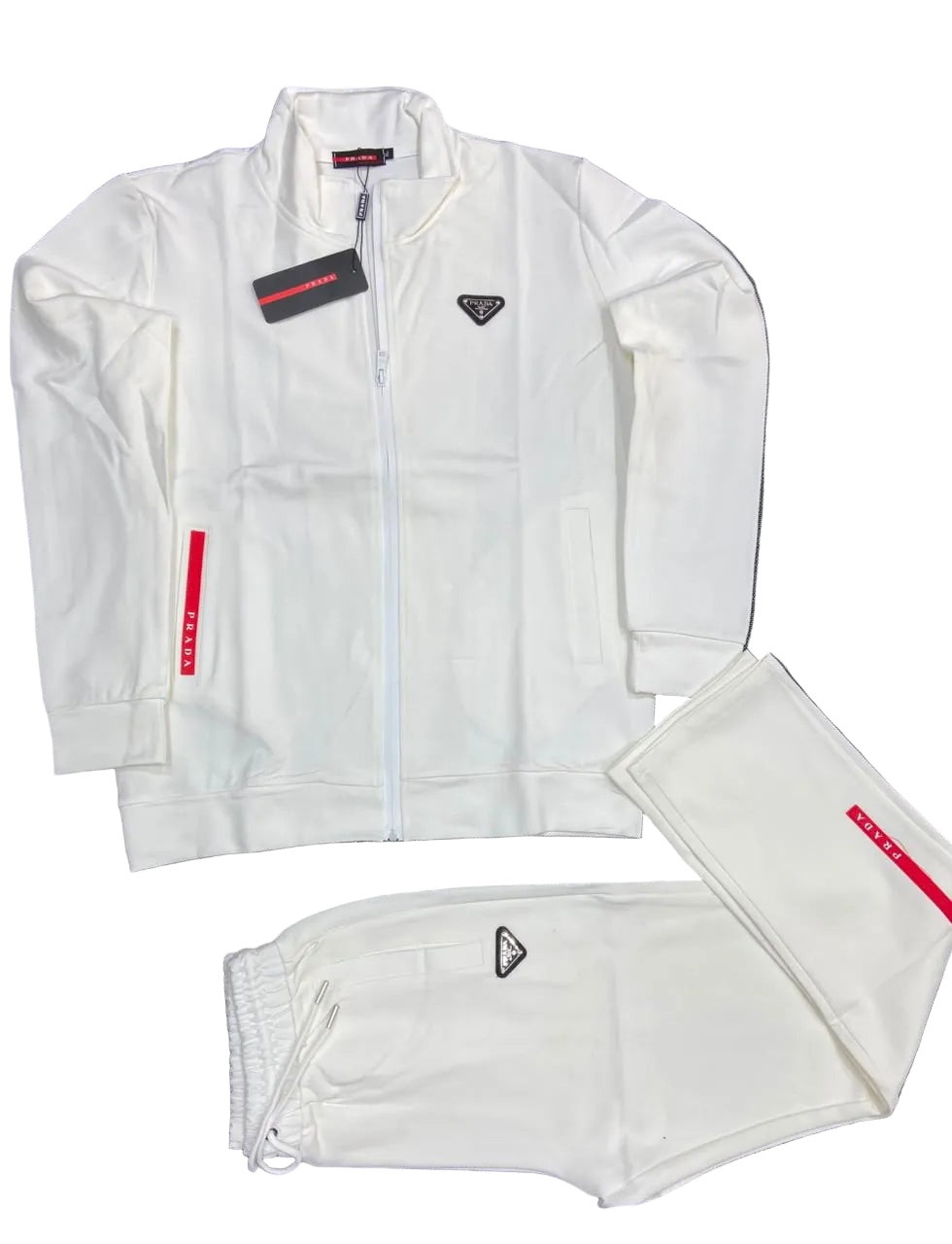 A white Prada tracksuit set laid flat on a dark gray textured background, including a zip-up jacket with a small black triangle logo on the chest, matching sweatpants with a drawstring waist, and folded pants showing a red Prada label.