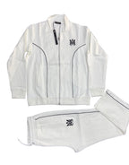 A white two-piece tracksuit set consisting of a zip-up jacket and matching shorts, laid flat on a dark gray textured background. The tracksuit features black piping details, a black embroidered 'MM' monogram logo on the left chest and left leg, and a black hangtag on the collar of the jacket.