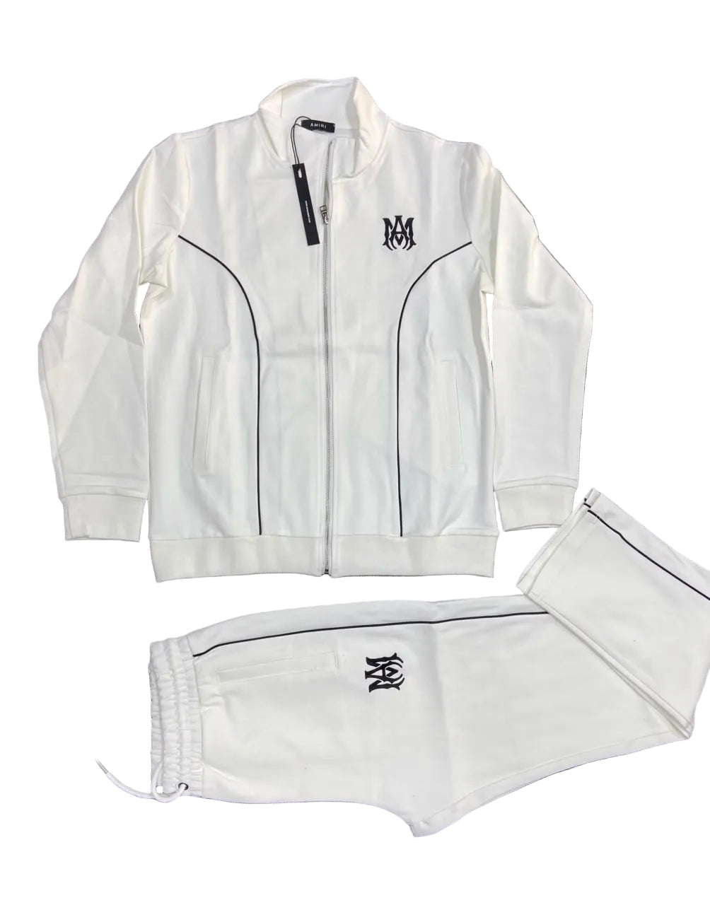 A white two-piece tracksuit set consisting of a zip-up jacket and matching shorts, laid flat on a dark gray textured background. The tracksuit features black piping details, a black embroidered 'MM' monogram logo on the left chest and left leg, and a black hangtag on the collar of the jacket.