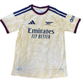 An Arsenal home soccer jersey by Adidas in a cream colorway, laid flat on a green turf background. The jersey features navy blue and burgundy trim, the Arsenal crest, the Adidas logo, and Emirates Fly Better sponsor branding.