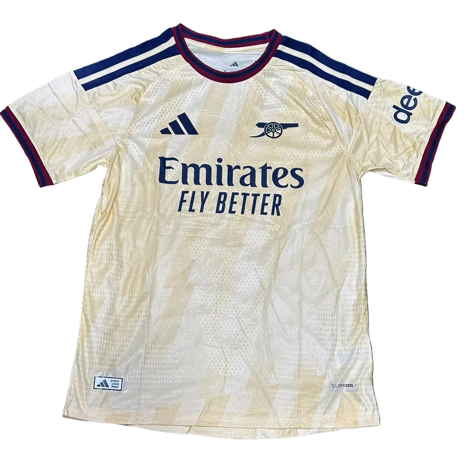 An Arsenal home soccer jersey by Adidas in a cream colorway, laid flat on a green turf background. The jersey features navy blue and burgundy trim, the Arsenal crest, the Adidas logo, and Emirates Fly Better sponsor branding.