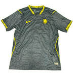 A black Nike Chelsea FC soccer jersey with yellow accents, laid flat on a green grass-like surface. The jersey features the Chelsea crest on the chest, a yellow Nike swoosh, and yellow graphic details on the collar, sleeve cuffs, and side panels.
