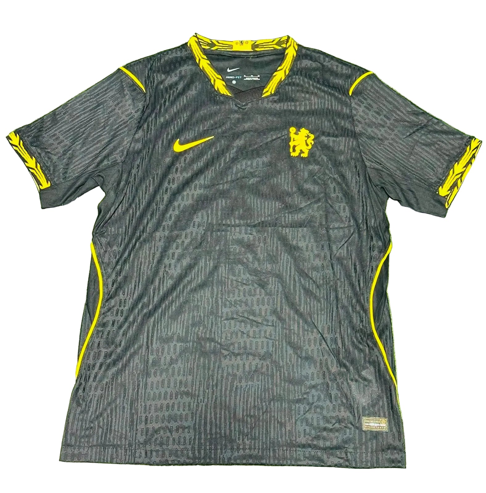 A black Nike Chelsea FC soccer jersey with yellow accents, laid flat on a green grass-like surface. The jersey features the Chelsea crest on the chest, a yellow Nike swoosh, and yellow graphic details on the collar, sleeve cuffs, and side panels.