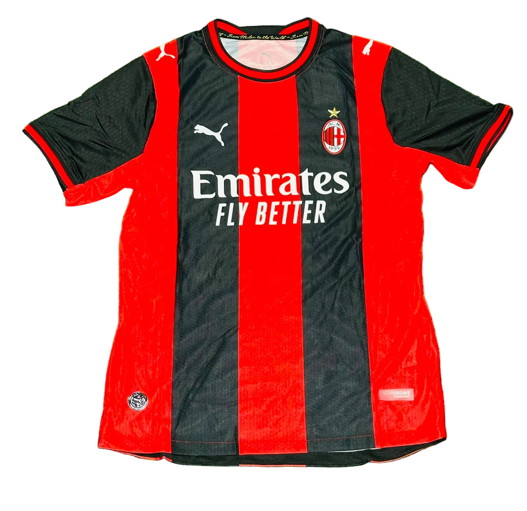 AC Milan home soccer jersey by Puma, featuring red and black vertical stripes with white sponsor and team logos, laid flat on green grass.
