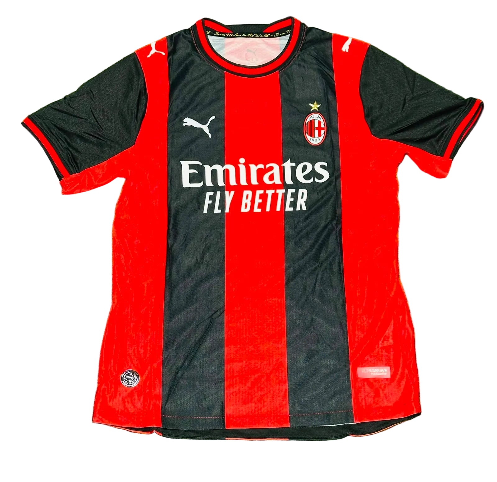AC Milan home soccer jersey by Puma, featuring red and black vertical stripes with white sponsor and team logos, laid flat on green grass.