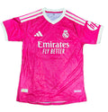 A pink Real Madrid soccer jersey with white accents, featuring the Adidas logo, Real Madrid crest, and Emirates Fly Better sponsor text, laid flat on a green grass background.