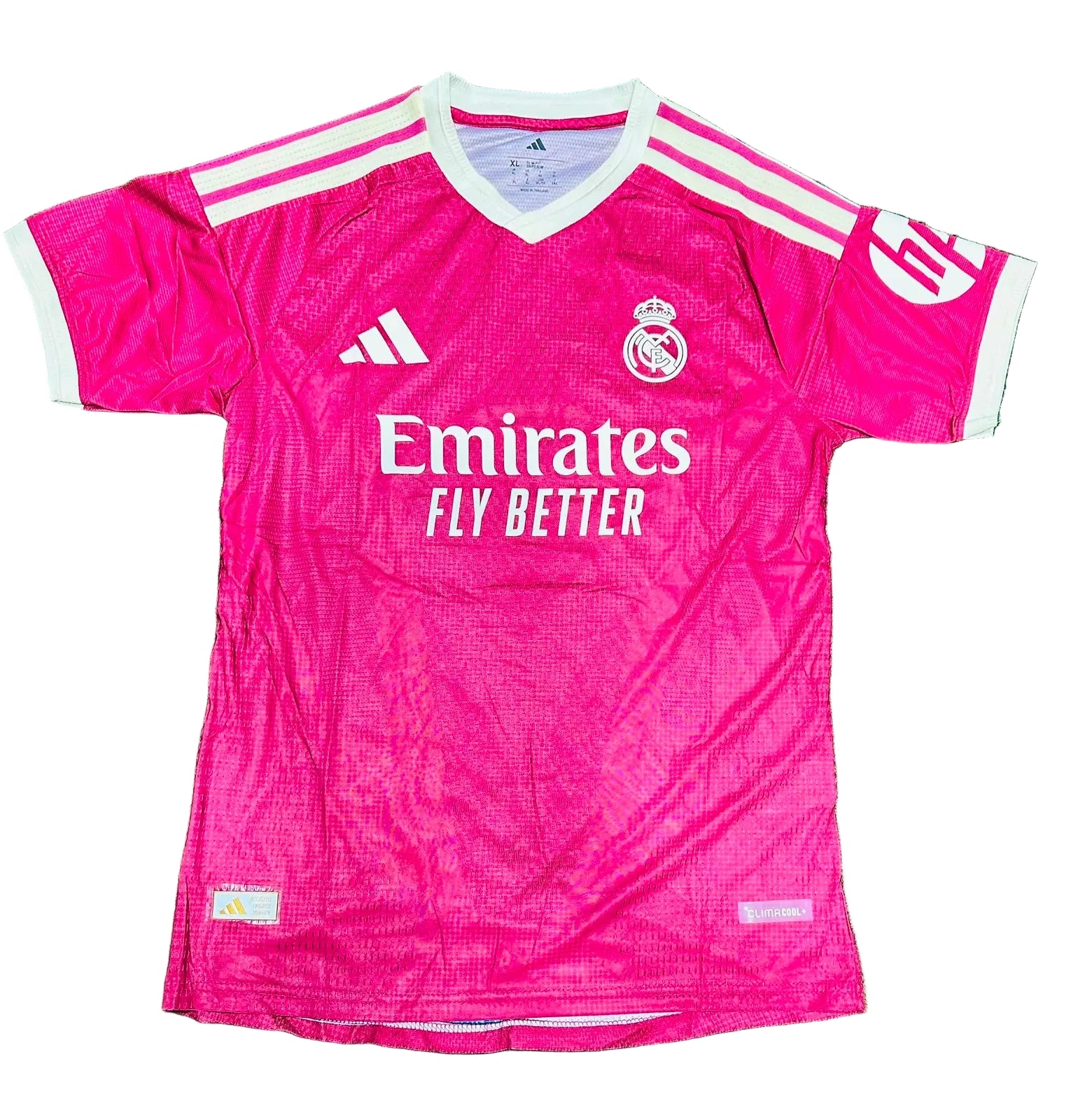 A pink Real Madrid soccer jersey with white accents, featuring the Adidas logo, Real Madrid crest, and Emirates Fly Better sponsor text, laid flat on a green grass background.