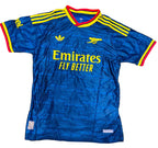 A blue Arsenal football jersey with yellow and red trim, laid flat on green grass. The jersey features the Adidas logo, Arsenal crest, Emirates Fly Better sponsor text, and yellow stripes on the shoulders.