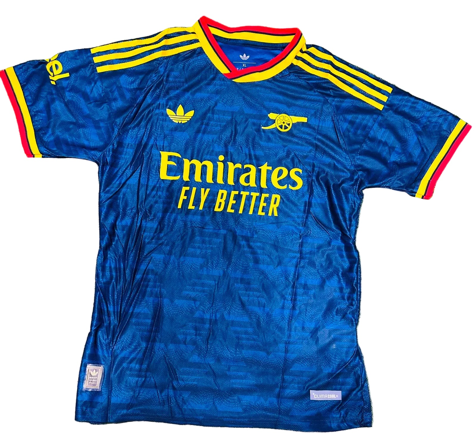 A blue Arsenal football jersey with yellow and red trim, laid flat on green grass. The jersey features the Adidas logo, Arsenal crest, Emirates Fly Better sponsor text, and yellow stripes on the shoulders.