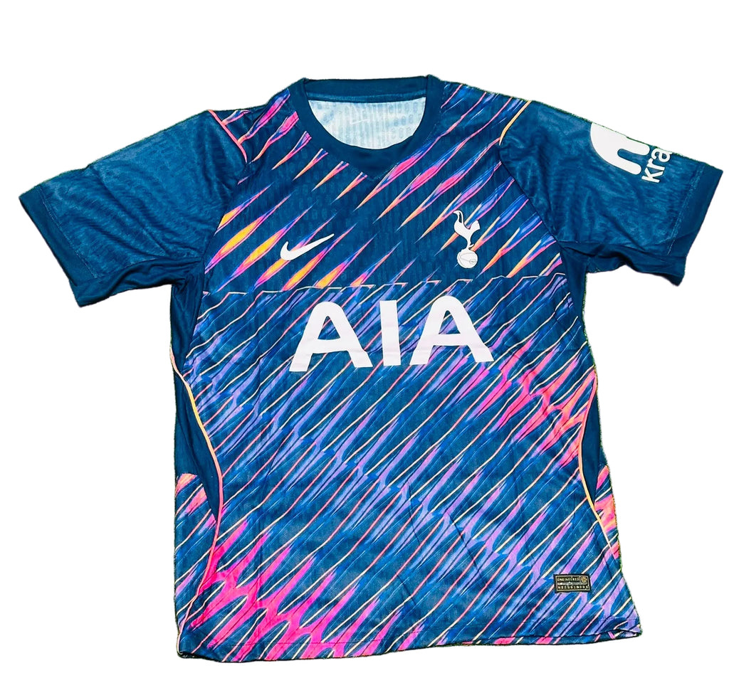 A blue Tottenham Hotspur soccer jersey with pink, orange, and yellow diagonal stripes, featuring the white AIA sponsor logo on the front, laid flat on green grass.