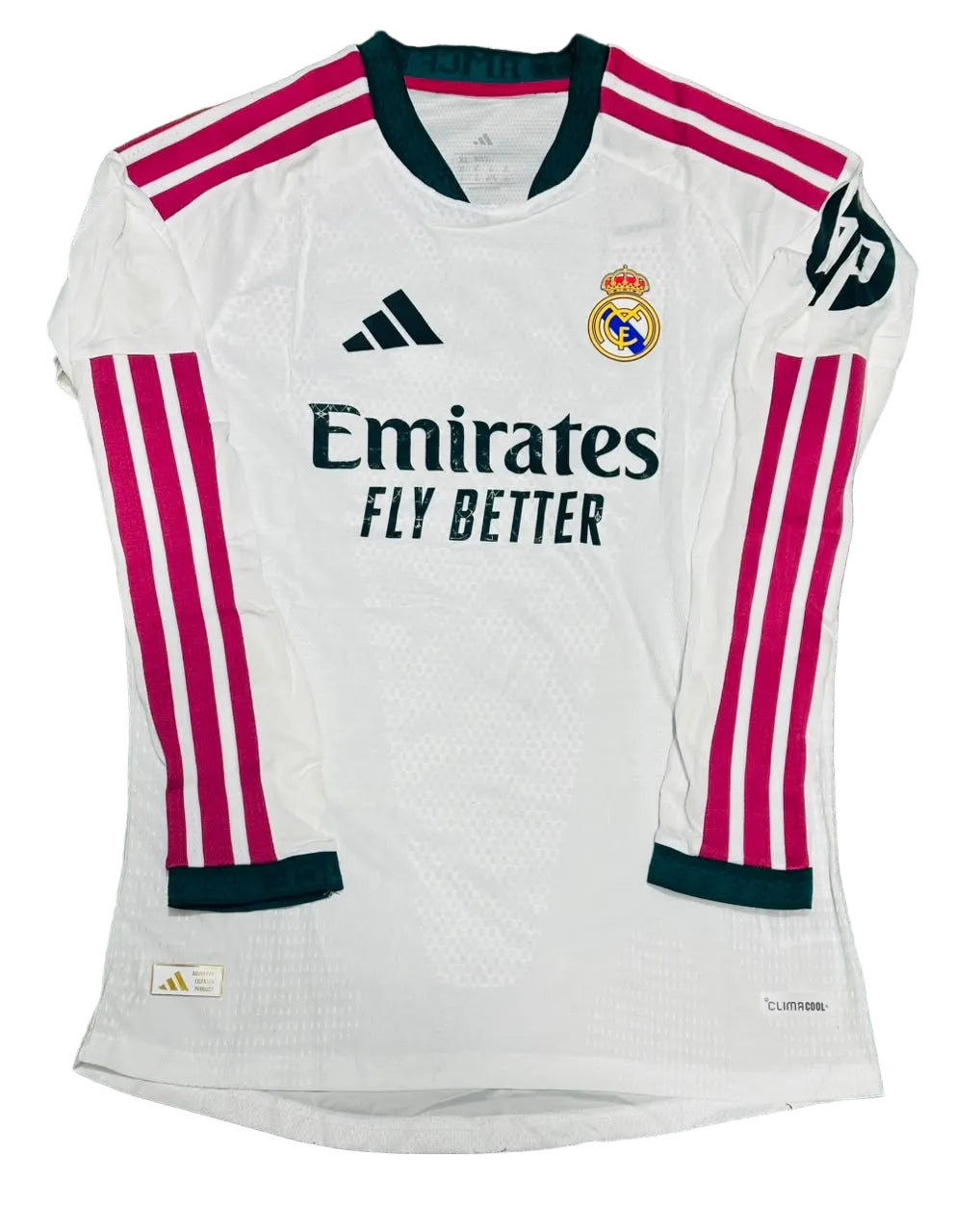 A white Real Madrid long-sleeve soccer jersey with pink and dark green accents, laid flat on a green grass background. The jersey features the Adidas logo, Real Madrid crest, Emirates Fly Better sponsor, and HP sleeve patch.