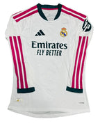 A white Real Madrid long-sleeve soccer jersey with pink and dark green accents, laid flat on a green grass background. The jersey features the Adidas logo, Real Madrid crest, Emirates Fly Better sponsor, and HP sleeve patch.