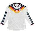 A white long-sleeve Germany soccer jersey with black, red, and yellow accents, laid flat on a green artificial turf background. The jersey features the Adidas logo, the German national team crest, and three-star embroidery on the chest.