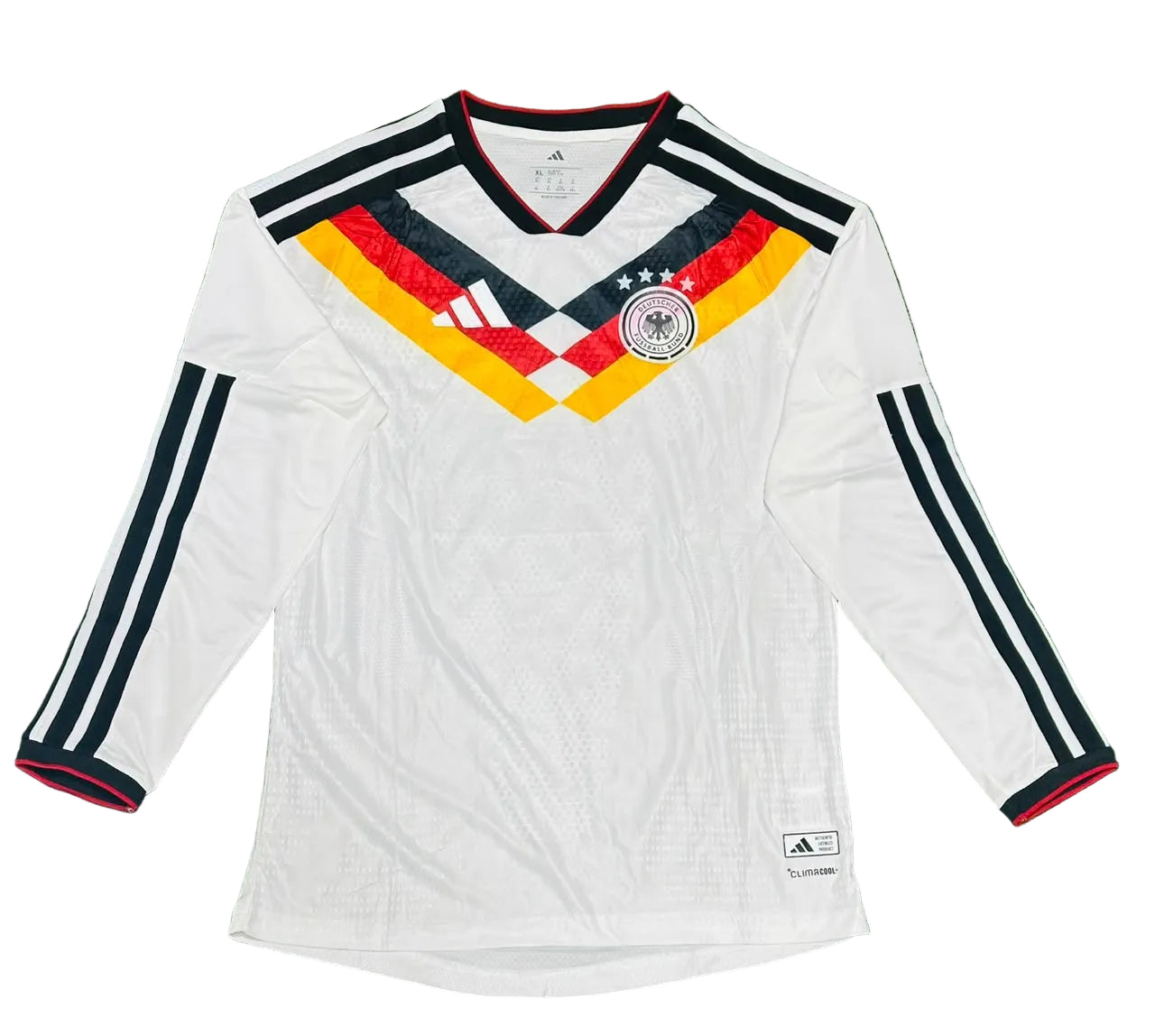 A white long-sleeve Germany soccer jersey with black, red, and yellow accents, laid flat on a green artificial turf background. The jersey features the Adidas logo, the German national team crest, and three-star embroidery on the chest.