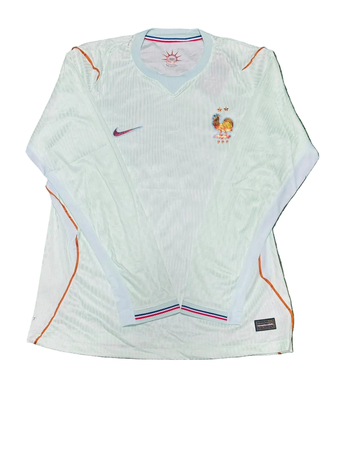 A white long-sleeve France national football team jersey by Nike, featuring orange piping, a blue collar, and the French crest on the chest, laid flat on green grass.