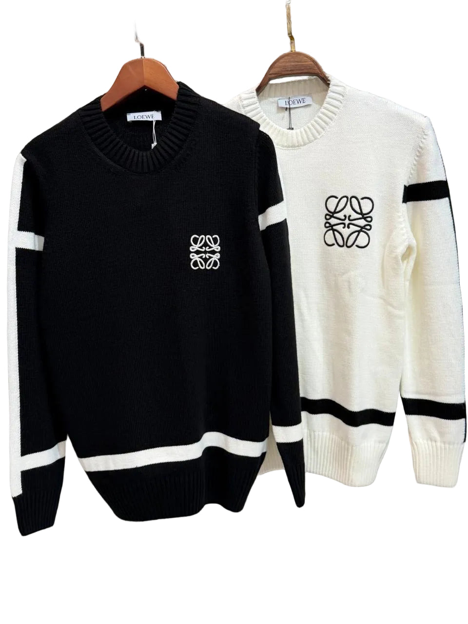 Two knit crewneck sweaters hanging on wooden hangers against a textured wall. The front sweater is black with white sleeve stripes, a white chest logo, and a white hem stripe.