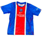 A Paris Saint-Germain soccer jersey laid flat on green grass. The jersey features a vertical color-blocked design with blue on the sides, a red center panel, and white stripes separating the sections.