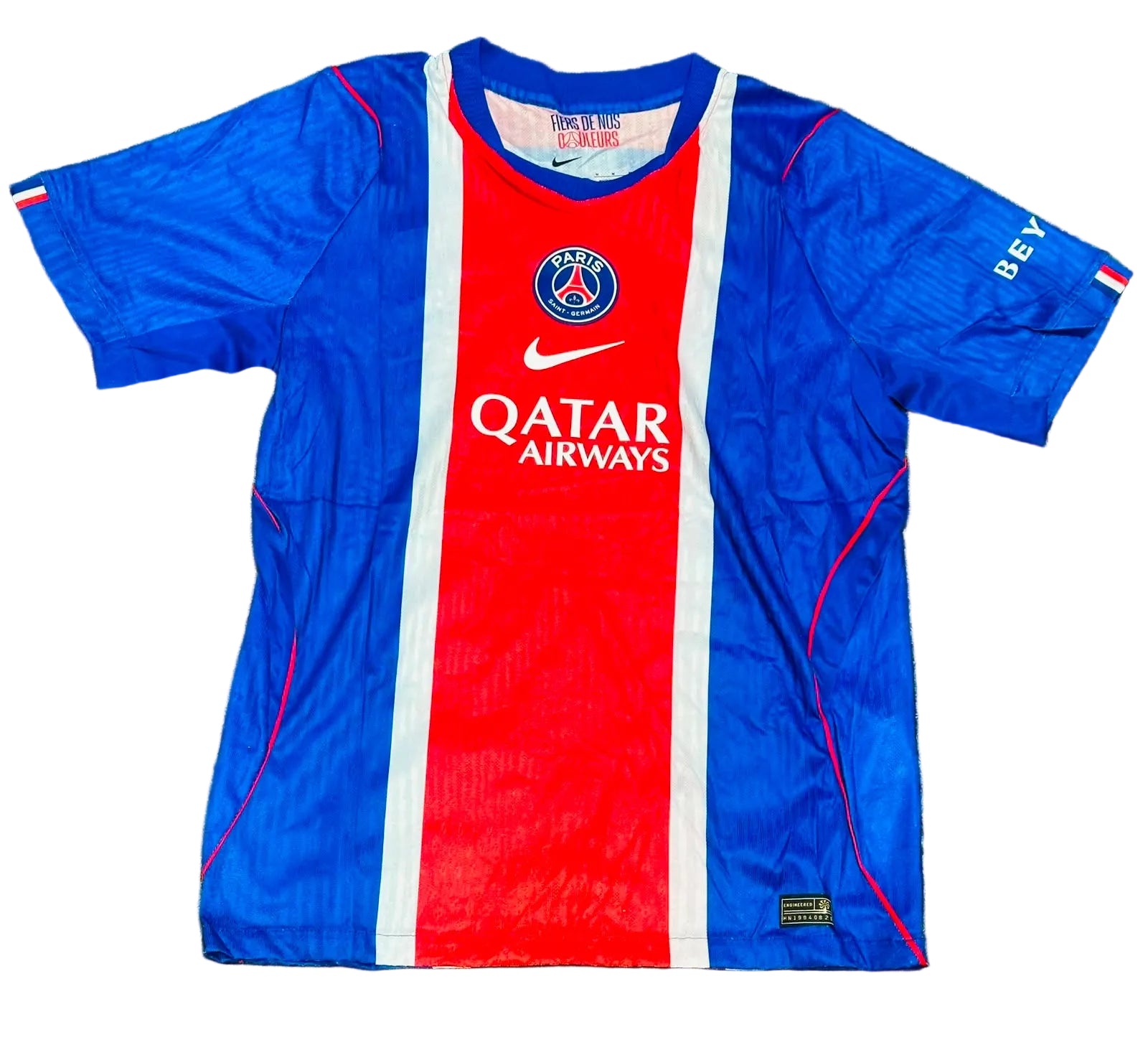 A Paris Saint-Germain soccer jersey laid flat on green grass. The jersey features a vertical color-blocked design with blue on the sides, a red center panel, and white stripes separating the sections.