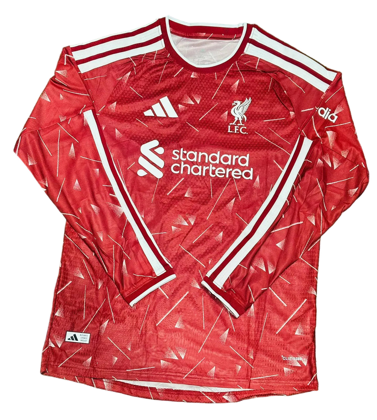 A red long-sleeve Liverpool football jersey with white stripes and logos, laid flat on green grass. Visible branding includes Adidas, LFC, and Standard Chartered.