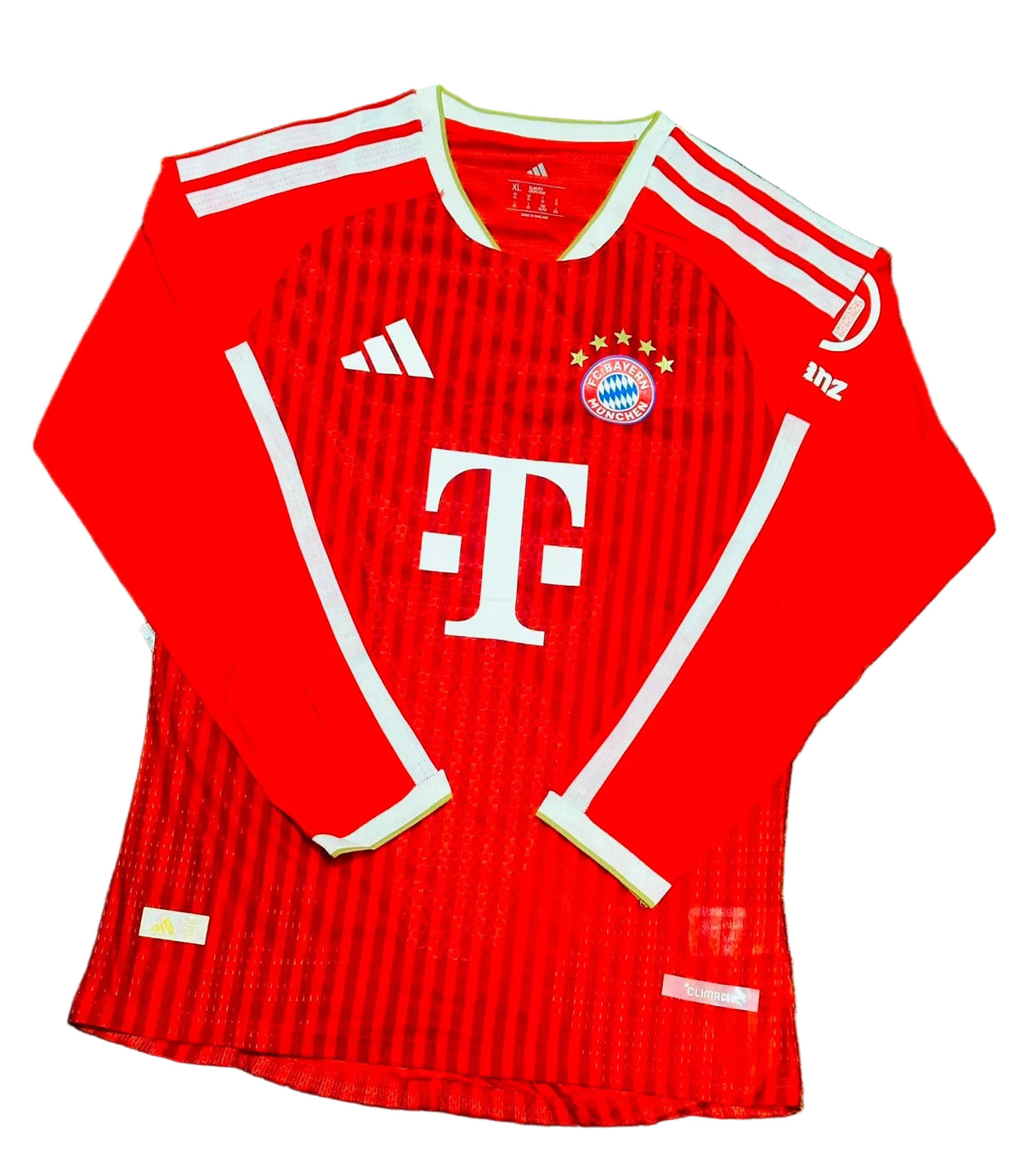 A red long-sleeve Bayern Munich soccer jersey with white stripes and logos, laid flat on green artificial turf. Visible branding includes the Adidas logo and the Bayern Munich club crest.